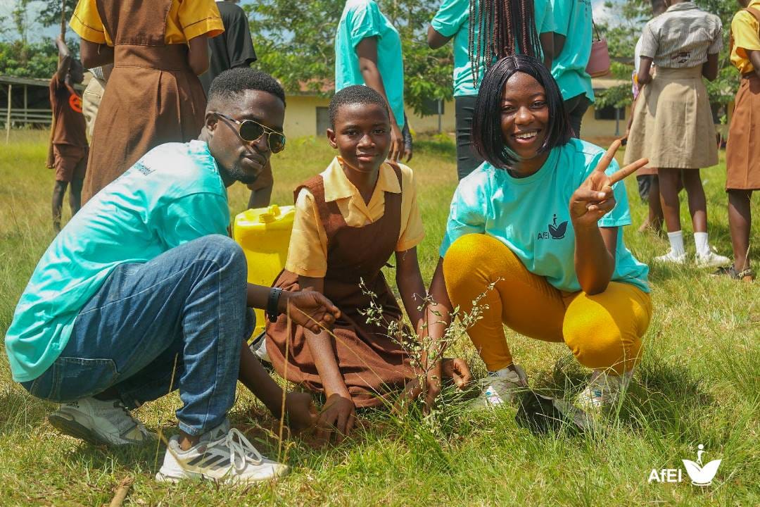 Two facilitators and a pupil crouching together to plant a seedling in grass, image 6 of 12