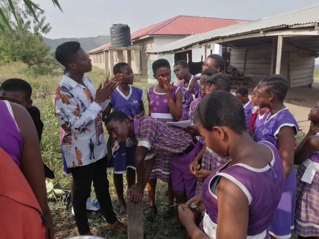 Pupils in purple school uniform gathered around a facilitator in a patterned shirt outdoors, image 10 of 12