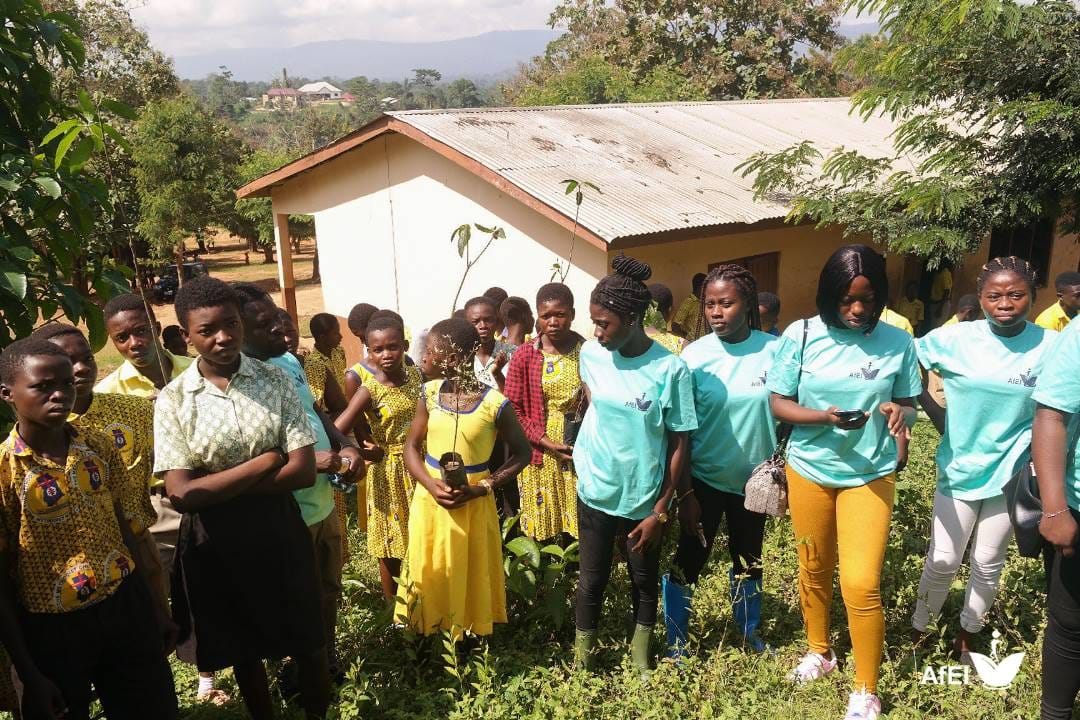 Students and facilitators in teal T-shirts gathered outdoors near a school building, several holding seedlings, image 4 of 12