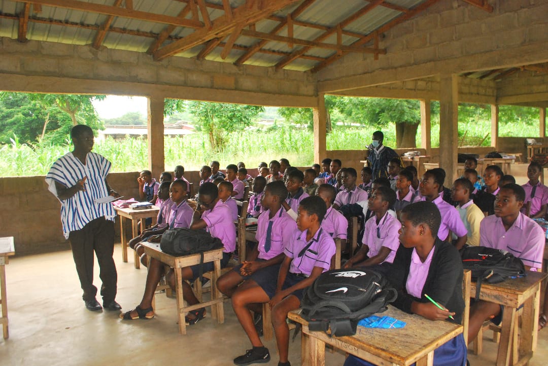 Students seated at desks in an open-sided classroom while a man in a smock speaks at the front, image 1 of 12