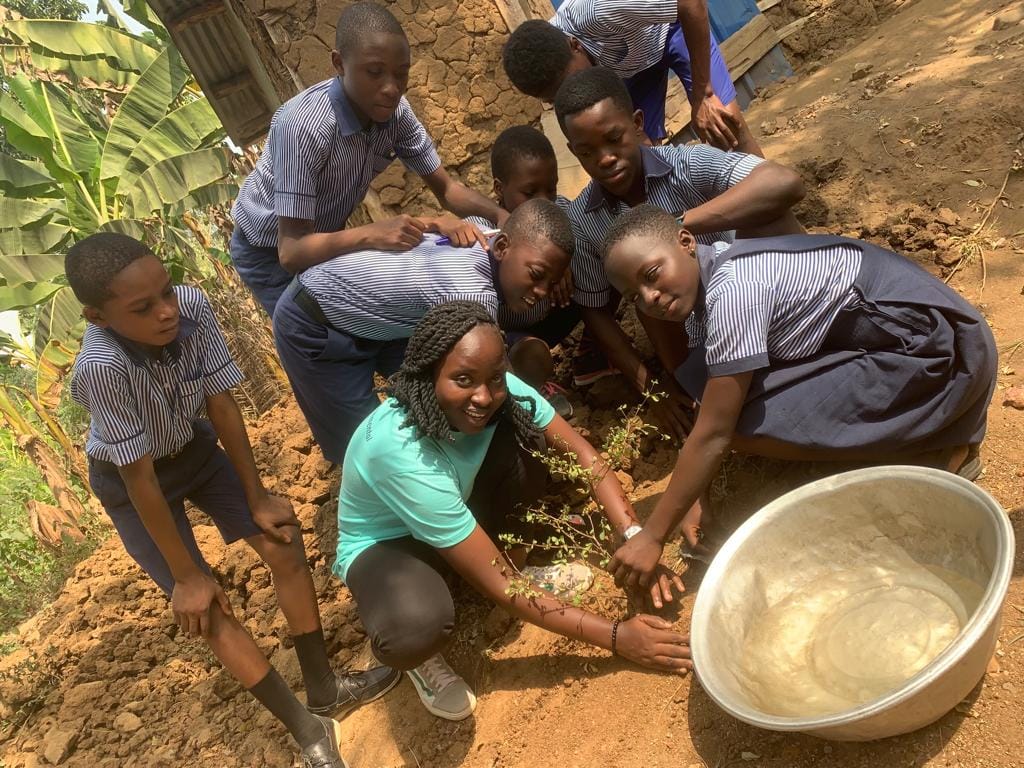 Pupils and a facilitator crouching around a planting hole with a basin of water beside them, image 5 of 14