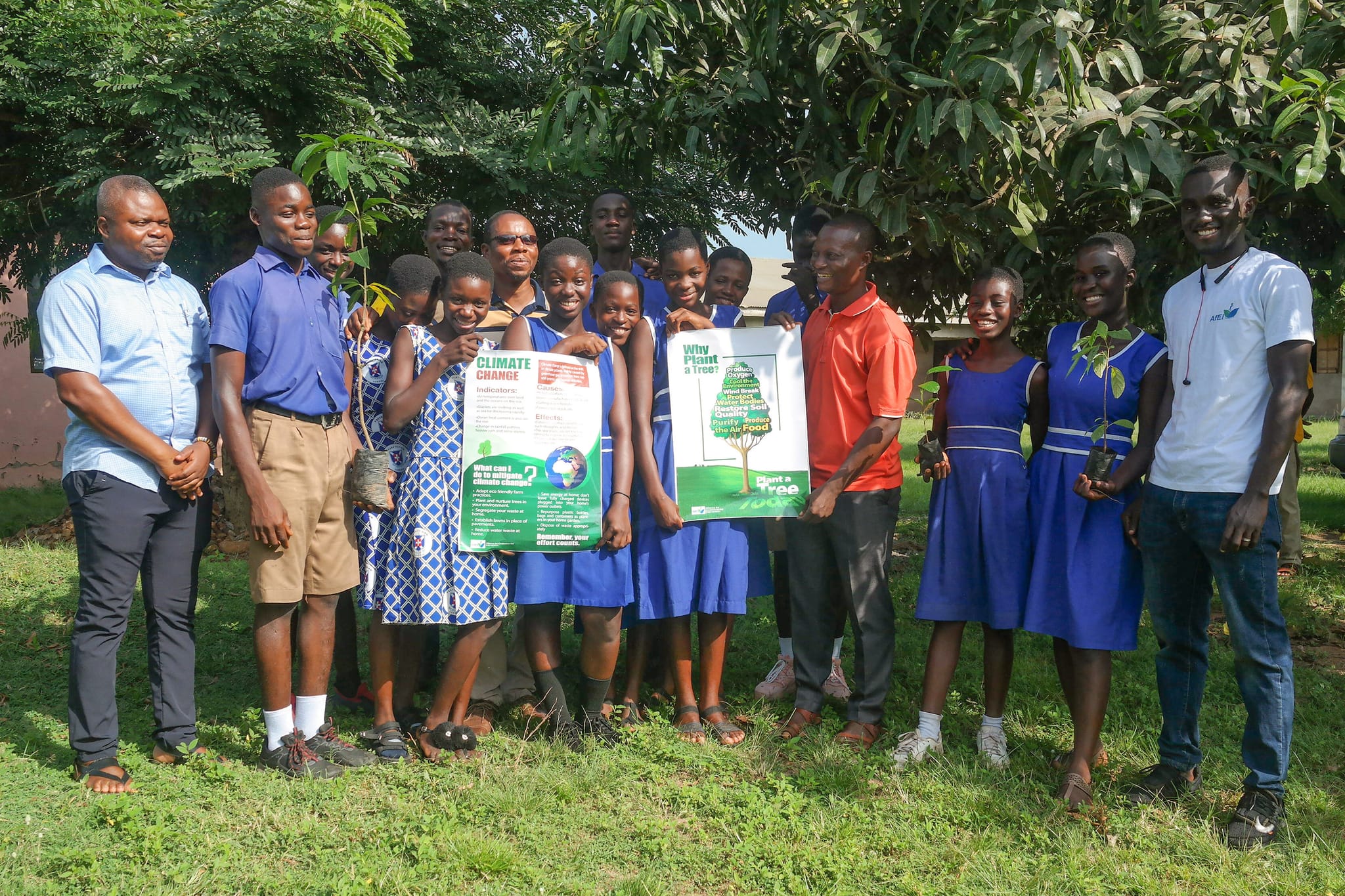 Pupils, teachers and facilitators posing outdoors, some holding tree-planting posters and seedlings, image 9 of 14