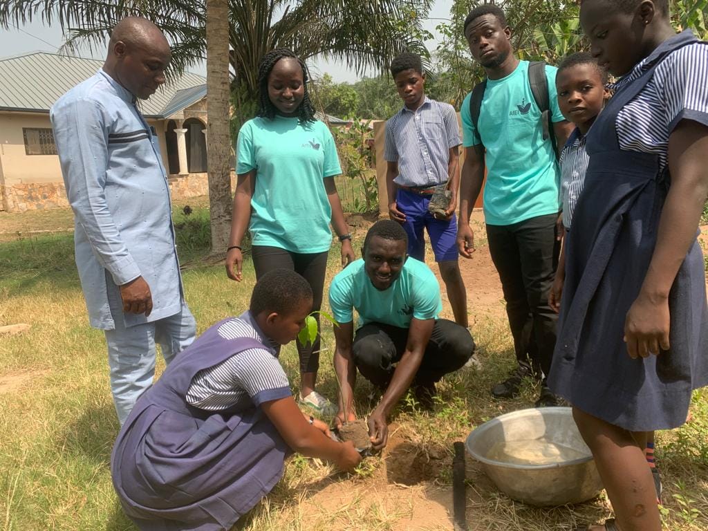 Pupils and facilitators gathered around a seedling being planted in the school compound, image 7 of 14